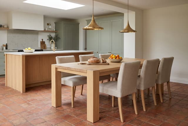 The Balkan Dining Table in an open-plan kitchen, sage cabinets and brass pendants, bread on the table