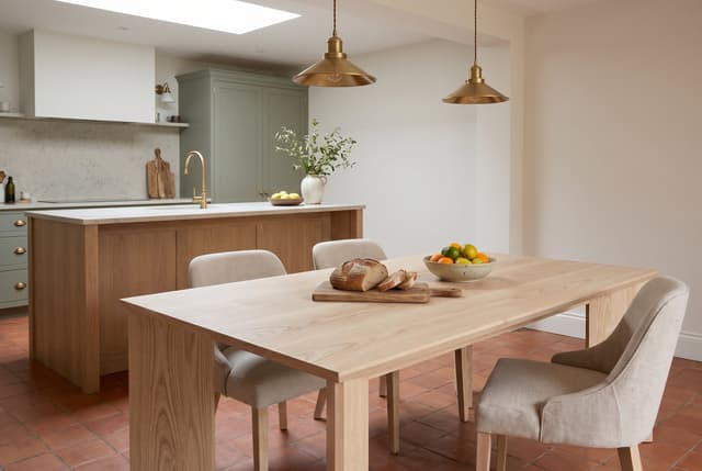 The Kouper Dining Table in an open-plan kitchen, sage cabinets and brass pendants, bread on the table