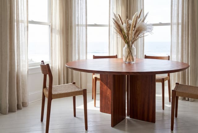 The Kouper Round Dining Table in a coastal bay window, sheer curtains and rope-seat chairs